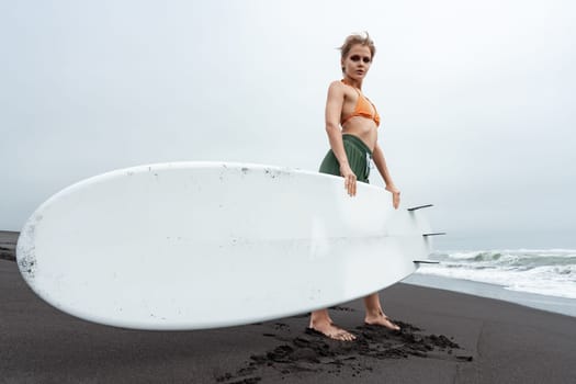 Female surfer standing on black sandy beach holding surfboard against background of sea waves during summer beach holiday. Woman looking at camera. Active lifestyle concept. Full length, wide shot