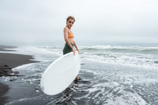 Woman surfer walking in ankle deep water of sea waves on beach carrying white surfboard against background of ocean during summer beach holiday. Sportswoman looking at camera. Full length, wide shot