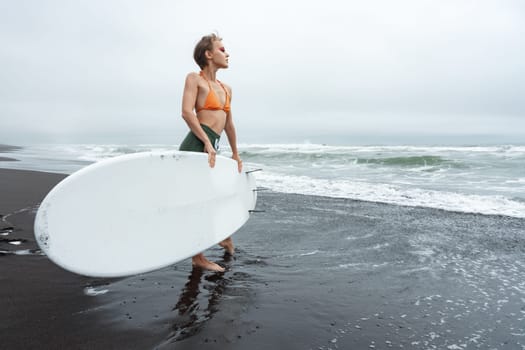 Attractive female surfer standing on black sandy beach holding white surfboard during summer beach holiday. This is perfect image to capture excitement and beauty of surfing. Full length, wide shot