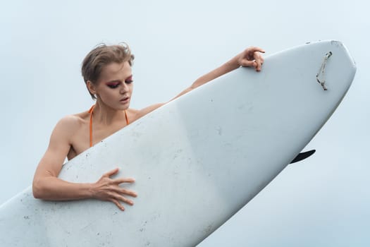 Female sports fashion model holding surfboard. She is standing behind surf board and looking down thoughtfully. Close-up view of beautiful woman surfer