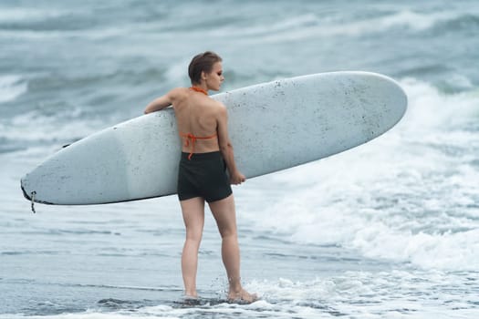 Woman surfer walking in ankle deep water on beach carrying white surfboard and looking at breaking waves of Pacific Ocean during summer sports training. Concepts of sport activities, healthy lifestyle