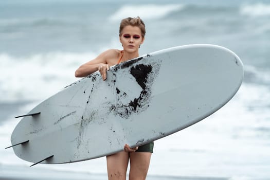 Woman surfer carrying white surfboard walking on beach and looking at camera on background of sea waves during summer vacation. Front view of sporting woman represents concept of physical activity