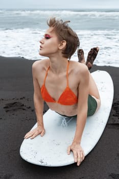 Woman surfer lying on front on surfboard, leaning on hands, arching back and crossing legs. Female sports fashion model looking away and warm up exercise before training during summer beach vacation