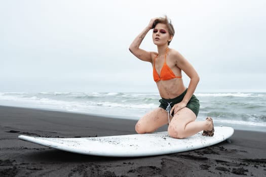 Woman surfer posing on her knees on white surfboard, looking at camera on black sand beach on background of ocean waves during summer holiday. Authenticity female surfer dressed in bikini top, shorts