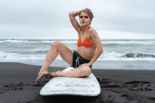 Woman surfer sitting on surfboard on black sand beach on background of ocean waves during summer holiday. Authenticity female athlete looking at camera. Sexy sportsperson in bikini top and shorts