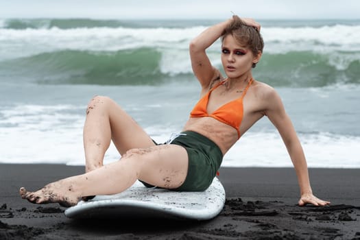 Female surfer sitting on surfboard, she looks so authentic and genuine, and even gazing right at camera. Sensuality woman rocking bikini top and shorts, and she looks like having time of her life