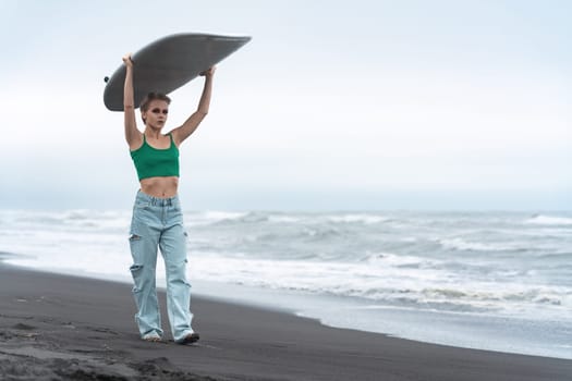 Woman surfer walking on black sandy beach, carrying white surfboard on head on background of sea waves during summer holiday. Hipster woman looking at camera, wearing in green top and blue jeans