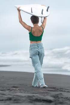 Rear view of female surfer is walking on black sandy beach with white surfboard on her head against background of ocean waves during solo travel summer. Hipster woman in green top and blue jeans