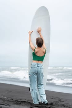 Rear view of woman surfer arms raised and holding surfboard vertically. Unrecognizable female standing on sandy beach, posing on background of ocean waves. Hipster woman wearing top and blue jeans