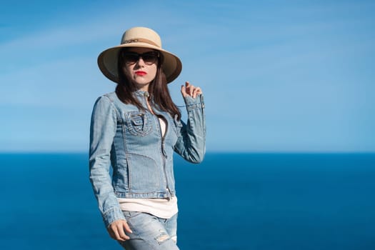 Portrait of woman in denim jacket, jeans, summer straw hat on head and sunglasses looking at camera. Hipster Caucasian ethnicity female standing against backdrop of blue sky and sea on sunny day