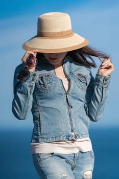 Portrait of female in denim jacket and jeans hiding with straw hat over his face. Hipster woman standing against backdrop of clear sky and blue sea on sunny day. Summer vacation concept