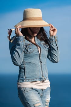 Portrait of hipster woman in denim jacket and jeans hiding with straw hat over his face. Adult female posing on background of blue cloudless sky and ocean on sunny day. Summer travel concept