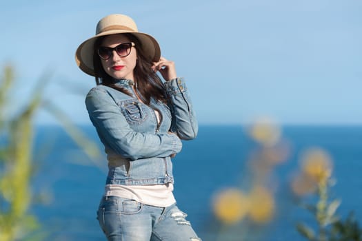Woman in sunglasses, straw hat, denim jacket and jeans pose beautifully against backdrop of blue sky and ocean. 40 year old hipster woman on vacation. Frame of green grass around edges of photo.