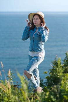 Full length portrait woman dressed in straw hat, denim jacket, jeans and boots standing on seashore in sunny weather. Hipster female raised hands, holding straw hat on head and looking into distance