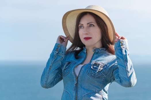 40 year old hipster woman raised hands and holding straw hat on her head. Caucasian ethnicity adult female in denim jacket posing against blue sky and ocean in sunny weather. Summer vacation concept