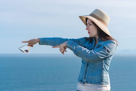 Hipster woman in straw hat and denim jacket shows with two hands direction to side. A serious 40 year old woman standing against backdrop of blue sky and ocean in sunny summer weather