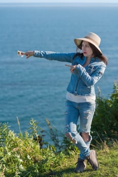 Full length serious woman in straw hat, denim jacket and jeans shows with two hands direction to side. Hipster woman standing cross legged against backdrop of blue sky and sea in sunny summer weather