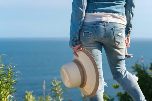 Cropped rear view below waist on legs of woman in blue jeans. Unrecognizable hipster female holding straw hat in one hand and sunglasses in other, standing in grass on seashore in sunny summer weather