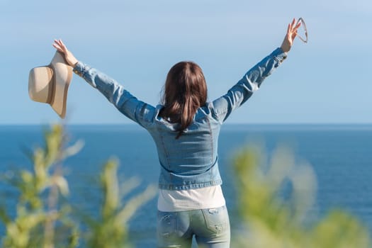 Rear view of unrecognizable woman raised hands, holding straw hat in one hand and sunglasses in other. Hipster female standing on seashore in sunny summer day against backdrop of blue sky and ocean