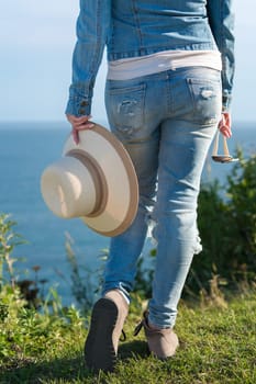 Rear view below waist on legs of hipster female in blue jeans and boots. Unrecognizable woman holding straw hat in one hand and sunglasses in other, standing in grass on seashore in sunny summer day