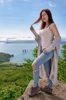 Adult Caucasian ethnicity woman in T-shirt, jeans, warm cardigan and boots stands on mountain shore against panoramic view of ocean on sunny summer day. Optimistic brunette woman looking at camera