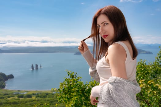 Adult Caucasian ethnicity woman in light T-shirt looking over shoulder at camera, stands on high mountain shore against panoramic view of green forest and ocean on sunny summer day