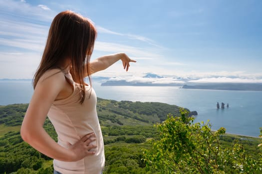 Faceless woman points with hand at blue sea below, standing high in mountains against panoramic view of green forest and ocean. Unrecognizable female tourist is resting on ocean in sunny summer day
