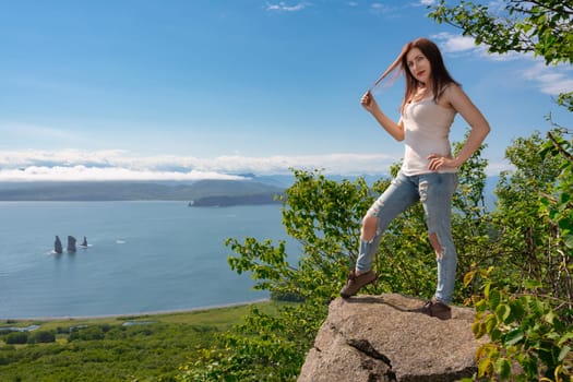 Adult woman in T-shirt, blue jeans and boots standing on a rocky shore in mountains against panoramic view of green forest and ocean on sunny summer day. Hipster female tourist looking at camera