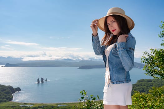 Adult hipster woman in straw hat, denim jacket and white shorts standing on high bank against backdrop of panoramic sea view on sunny summer day. Copy space