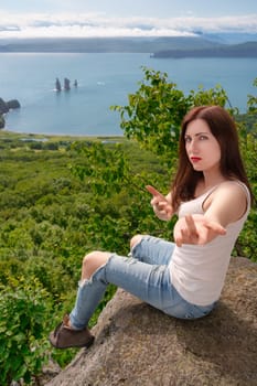 Bossy woman in T-shirt and blue jeans sitting on cliff on rocky shore in mountains against panoramic view of green forest and ocean on summer day. Female pulls hands into camera, inviting to travel