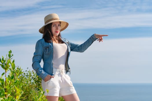 Hipster woman looking at camera and pointing one hand to side. Female dressed in straw hat, denim jacket, white shorts and standing on high bank against background of blue sky and strip of sea below