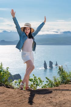 Hipster woman looking at camera with her hands raised, dressed in straw hat, denim jacket, white shorts and sandals, stands on high mountain against background of ocean on sunny summer day