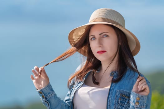 Mysterious adult woman with gray eyes looking at camera. Hipster dressed in straw hat, denim jacket and light T-shirt. Female tourist standing on blurred natural background of sea and forest