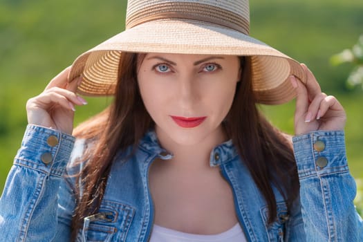 Portrait of woman with gray eyes looking at camera. Female hipster standing on blurred green natural background of forest. Front view of authentic brunette woman dressed in straw hat and denim jacket