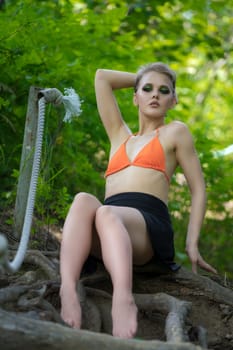 Sensuality woman sitting and poses in forest on ladder made of tree roots. Caucasian adult female dressed in orange bikini top, mini skirt. Blonde model with short hair and make-up, looking at camera
