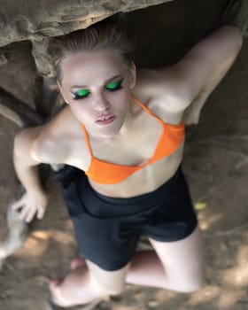 High angle shot of blonde woman with eyes closed relaxation on ground on roots of trees in summer forest. Model with bright make-up and short hair. Sexy female dressed in orange bikini top, mini skirt