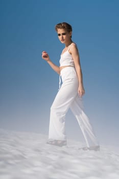 Caucasian ethnicity female tourist walking through snowfield with light fog on sunny day with blue sky. Sensual blond adult woman wearing in white cropped top, white pants and sandals