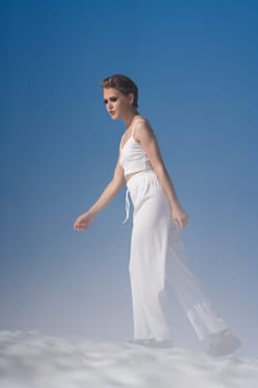 Beautiful blond woman dressed in white crop top, white pants and sandals. European female tourist walking through snow with light fog on sunny day with blue sky. Full length, side view