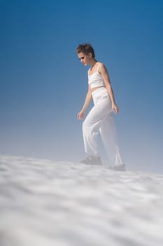 Woman walks through snowfield with light fog on sunny day with blue sky. Elegant blond adult woman dressed in white crop top, white pants and sandals