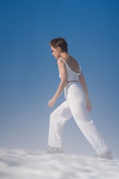 Slim blond woman walks through snow with light fog on sunny weather with blue sky. Pretty adult female with short hair in white crop top and white pants, sandals. Full length, side view