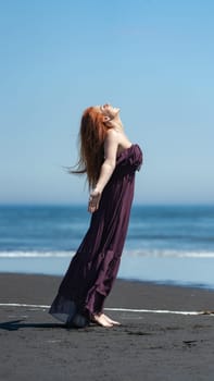 Happy redhead woman in purple brown long dress stands on sandy beach with head looking upward and eyes closed, arms to side on background blue sea on summer day with clear sky. Full length, side view