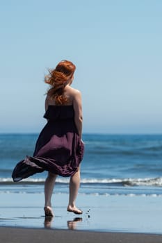 Rear view of woman in dark purple long dress, strolling barefoot on sandy beach towards crystal-clear water of blue sea on sunny day with clear sky. full-length view of female is simply breathtaking