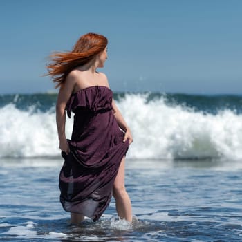 Redhead woman in long dress with knee high hem standing ankle deep in water on beach and turned head, looking over shoulder to sea in anticipation of crashing waves during summer holidays on sunny day