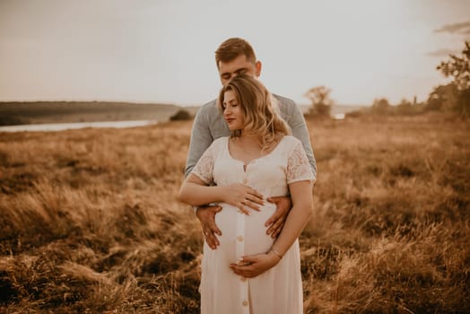 husband hugs pregnant wife keep his hands on big round tummy. Happy family resting in nature hugs kisses in summer at sunset. future mother Caucasian woman in white cotton dress father in shorts