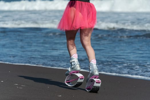 KAMCHATKA, RUSSIA - JUNE 15, 2022: Sporty woman legs in Kangoo Jumps sports boots, black swimsuit and short skirt running and jumping on beach during outdoors aerobic workout. Rear view, crop shot