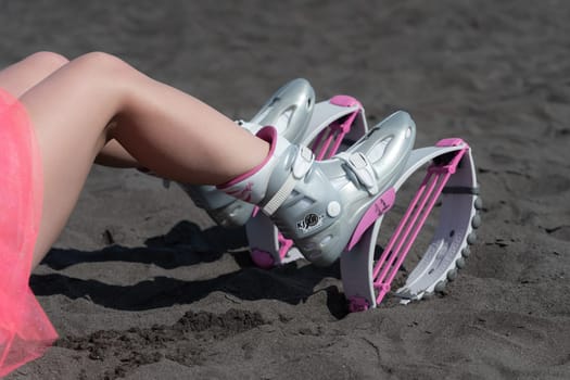 KAMCHATKA, RUSSIA - JUNE 15, 2022: Closeup slim female legs in sports Kangoo Jumps boots, short pink skirt on black sandy beach before outdoor aerobic fitness workout. High angle shot of trendy boots