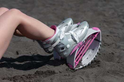 KAMCHATKA, RUSSIA - JUNE 15, 2022: Closeup slim female legs in sports Kangoo Jumps boots on black sandy beach before outdoor aerobic exercising, fitness workout. High angle shot trendy jumping boots