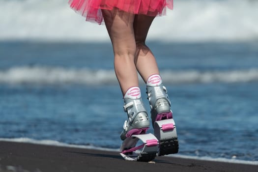 KAMCHATKA, RUSSIA - JUNE 15, 2022: Rear cropped view of sportive woman legs in sports boots Kangoo Jumps and short pink skirt jumping and running on beach during aerobic training session outdoors