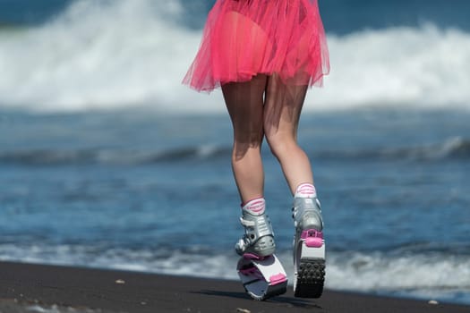 KAMCHATKA, RUSSIA - JUNE 15, 2022: Rear view of fit female legs in short pink skirt and sports jumping boots Kangoo Jumps running on sandy beach during training fitness session. Low section, crop view