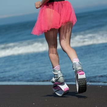 KAMCHATKA, RUSSIA - JUNE 15, 2022: Rear view of female legs in sports Kangoo Jumps boots, swimsuit and short pink skirt running and jumping on sandy beach during fitness training. Dutch angle shot